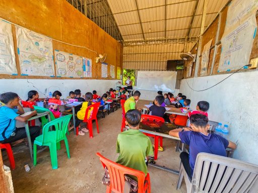 Kinder sitzen in einem Klassenraum, schreiben an Tischen mit bunten Stühlen.