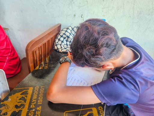 Ein junger Mensch schreibt mit dem Kopf auf einem Tisch, umgeben von verschiedenen Gegenständen.